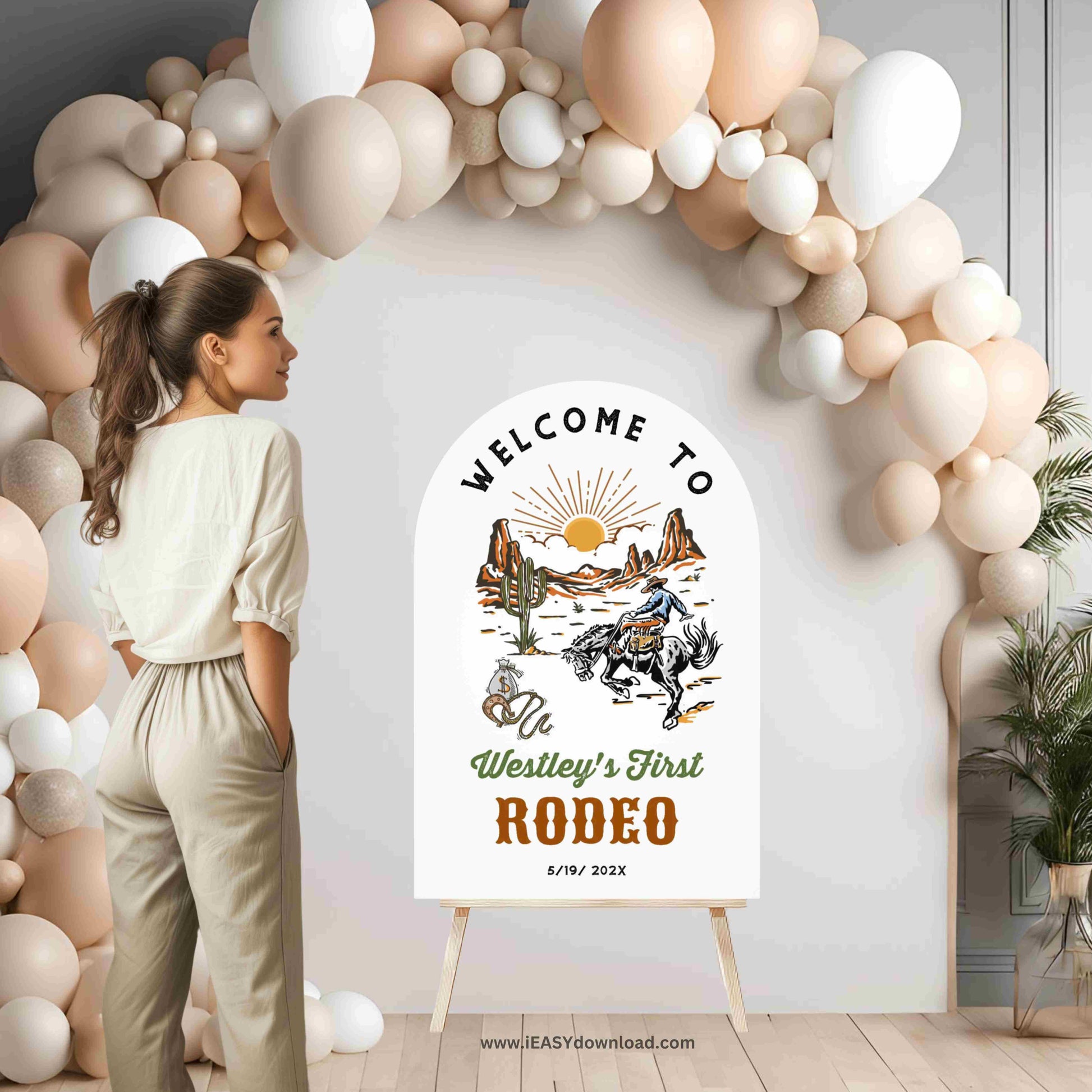 Woman standing in front of a 'Welcome to Westley's First Rodeo' sign with a balloon arch.