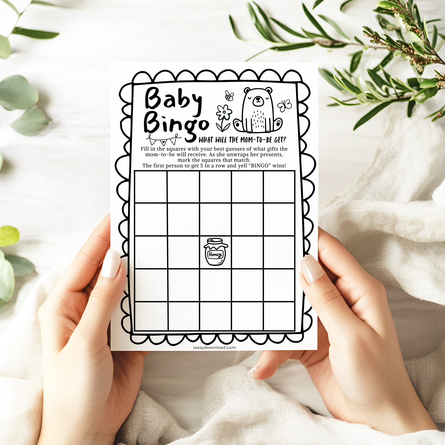 Baby Bingo game card held by hands with a white background and greenery.