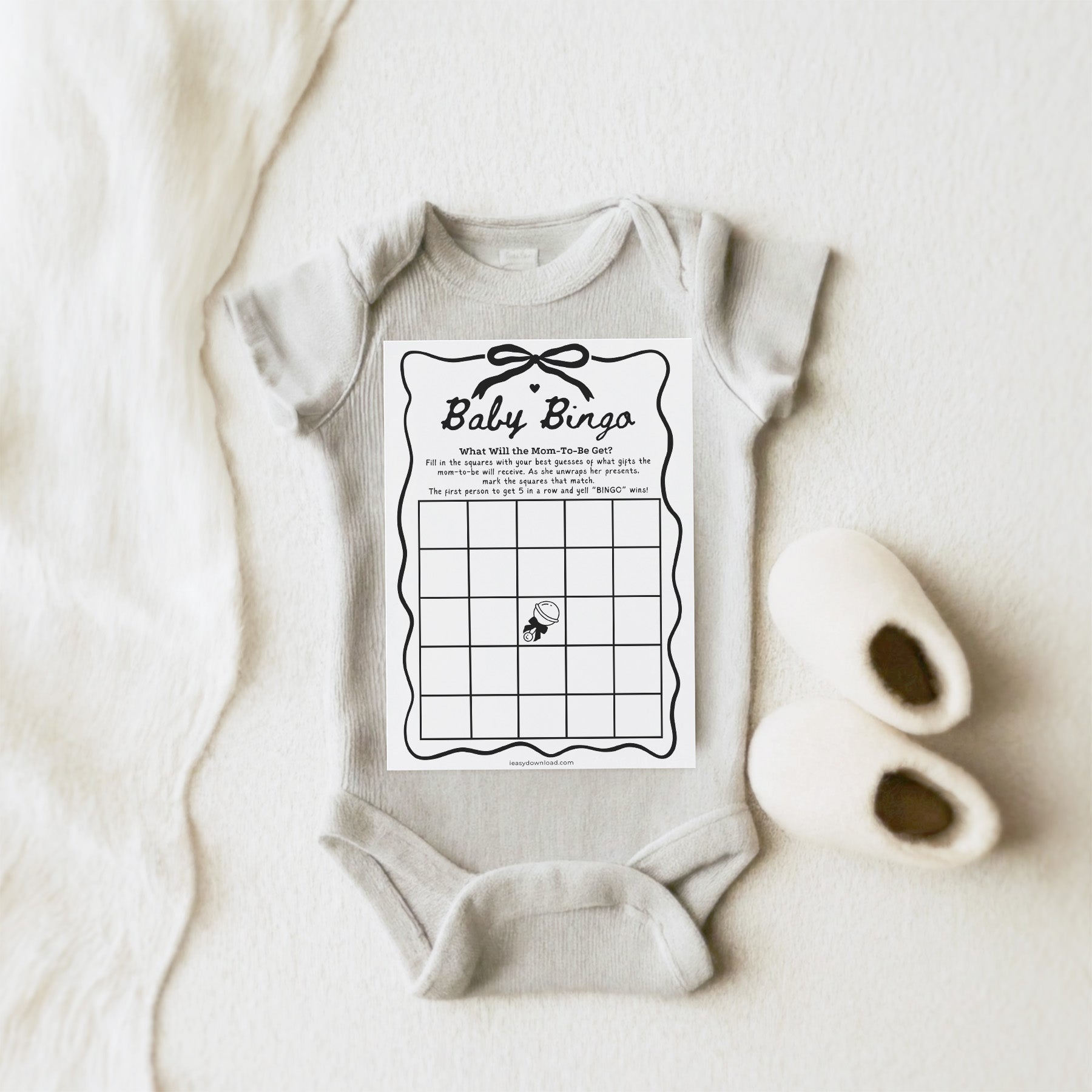 Baby onesie with Baby Bingo game card printed on it, placed on a light fabric background.